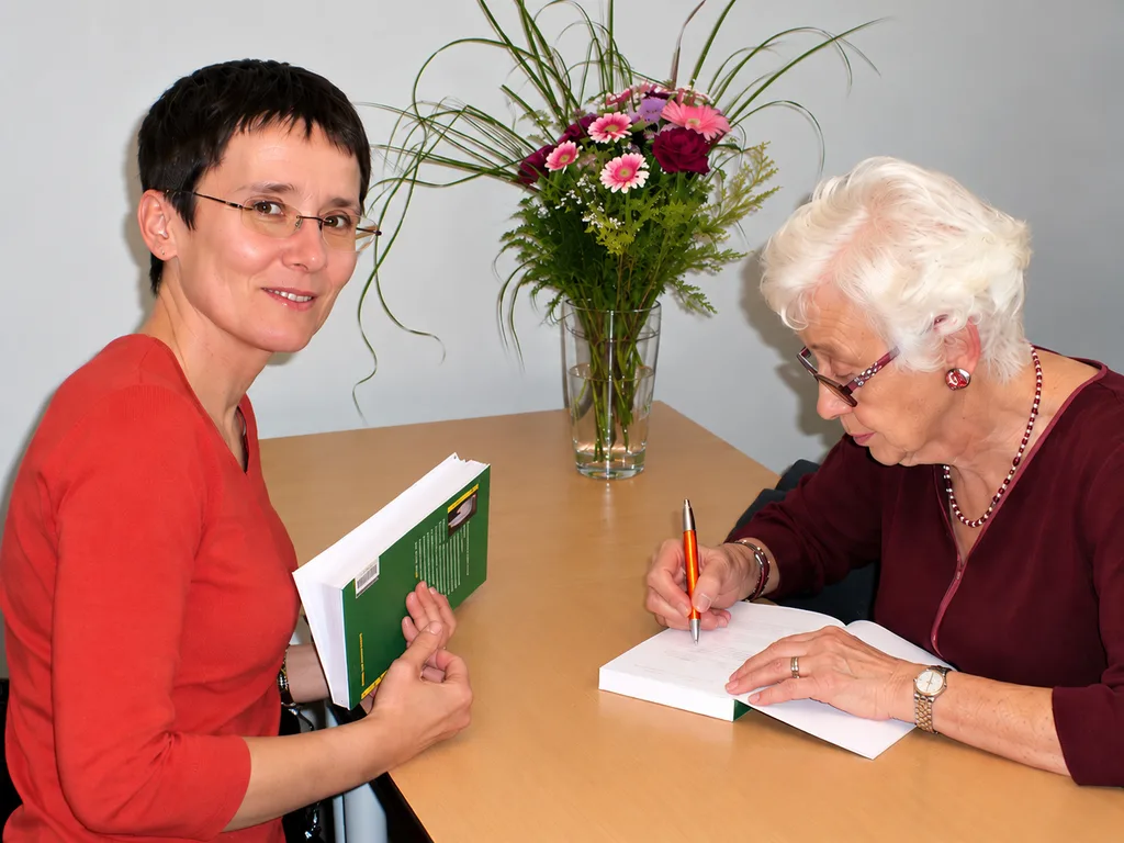 Andrea Stix und Naomi Feil sitzen an einem Tisch mit Büchern und einem Blumenstrauß